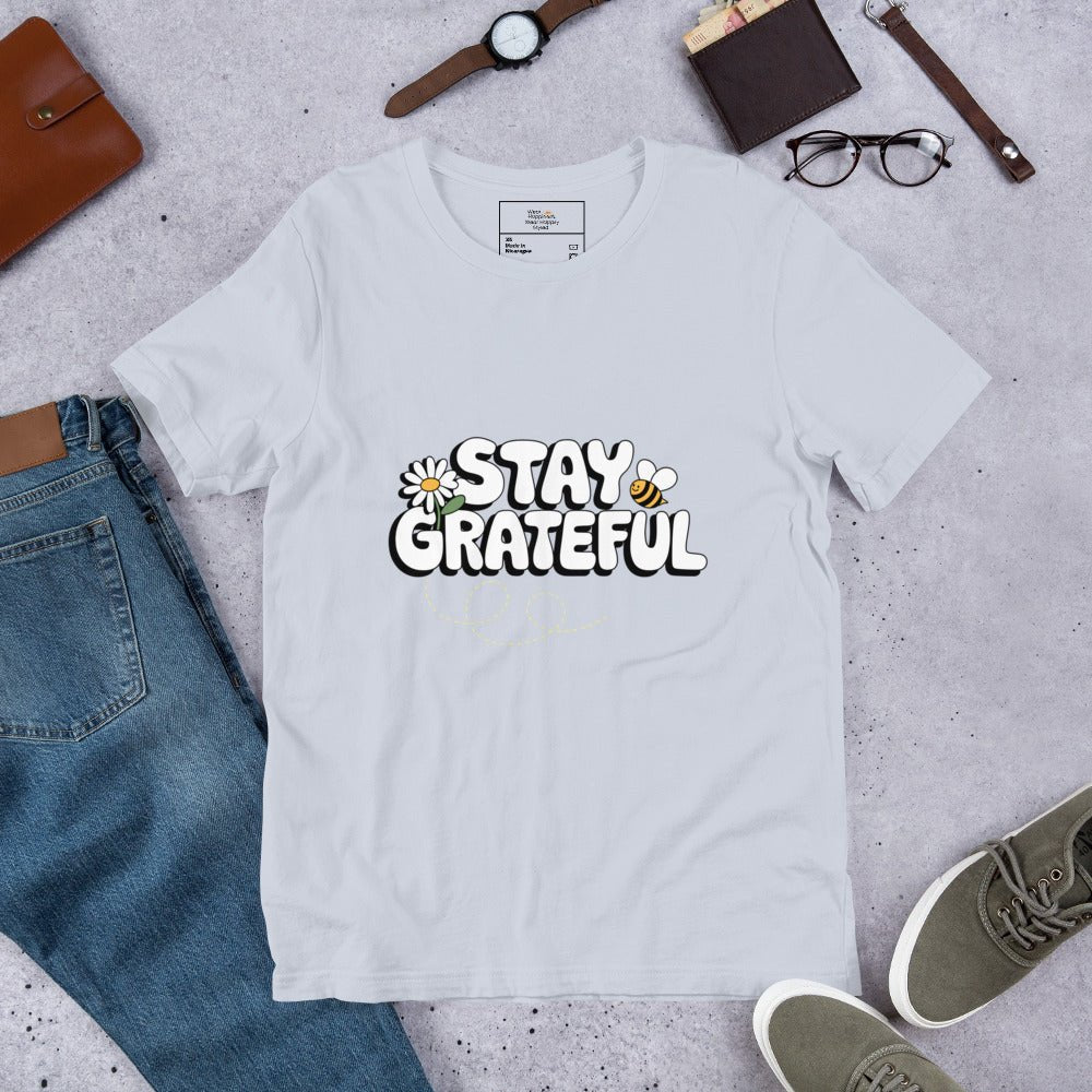 Light blue t-shirt with “Stay Grateful” text design, featuring daisies and a bee trail, styled flat with jeans, sneakers, glasses, a watch, and leather accessories on a gray background.