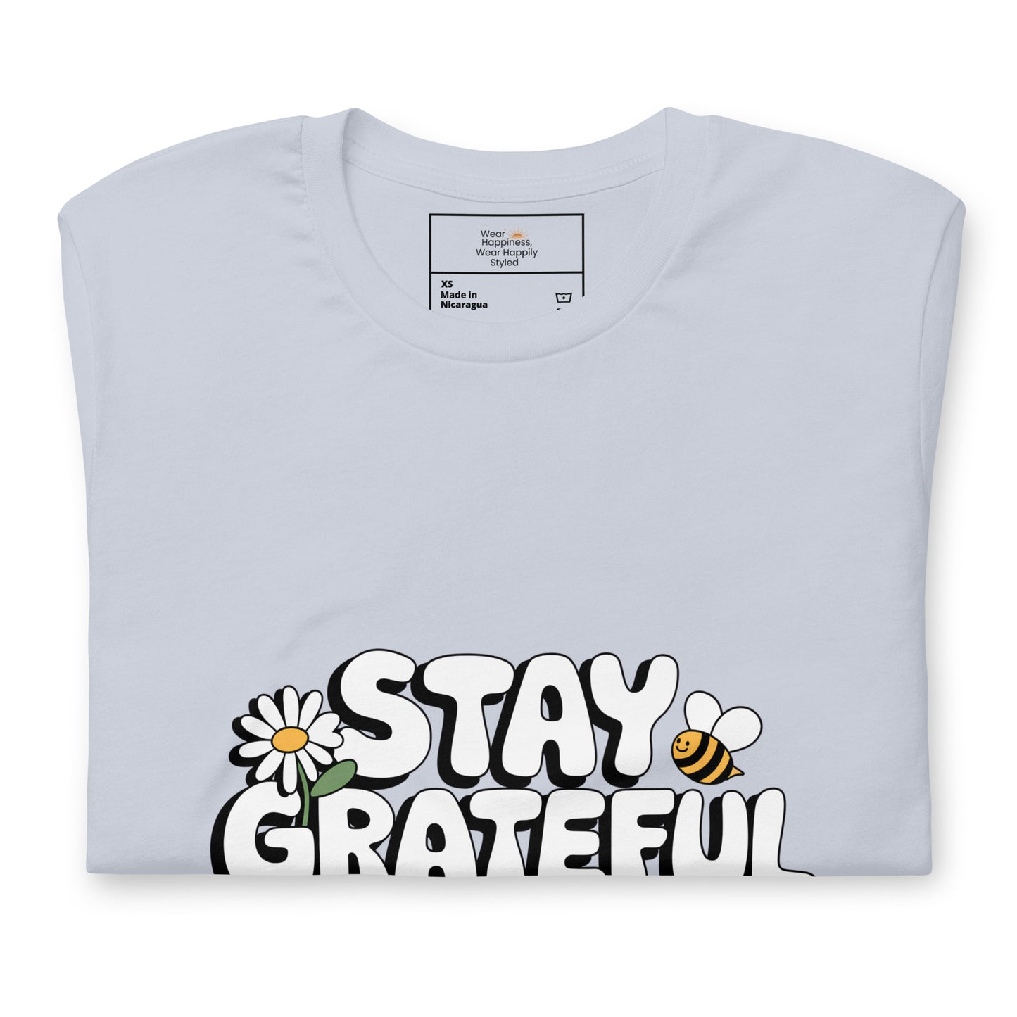 Folded light blue t-shirt with a bold white “Stay Grateful” design, featuring a daisy flower and a bee illustration, displayed on a white background.
