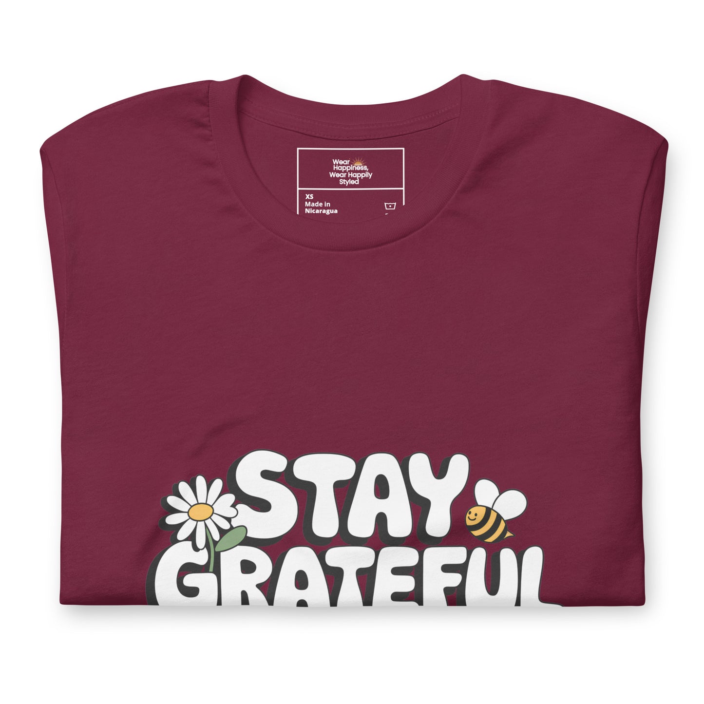 Folded maroon red t-shirt with a bold white “Stay Grateful” design, featuring a daisy flower and a bee illustration, displayed on a white background.