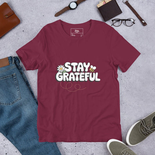 Maroon red t-shirt with “Stay Grateful” text design, featuring daisies and a bee trail, styled flat with jeans, sneakers, glasses, a watch, and leather accessories on a gray background.