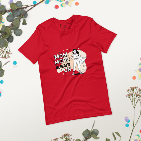 Red t-shirt with 'Mom Mode: Always On' superhero mom design, laid flat on a white surface with eucalyptus leaves and colorful confetti for decoration."