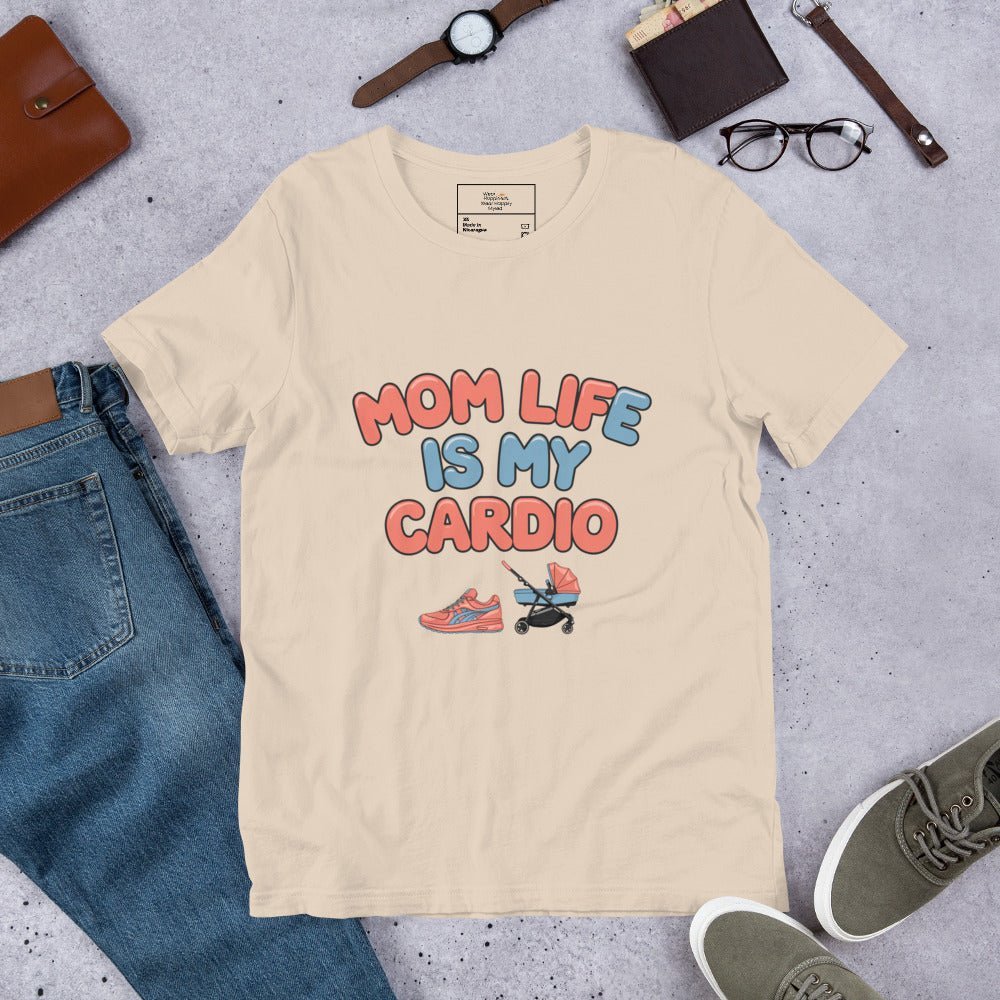 Soft cream unisex t-shirt laid flat on a surface next to jeans and accessories, featuring the phrase 'Mom Life Is My Cardio' in pastel letters with sneakers and pacifier graphic.