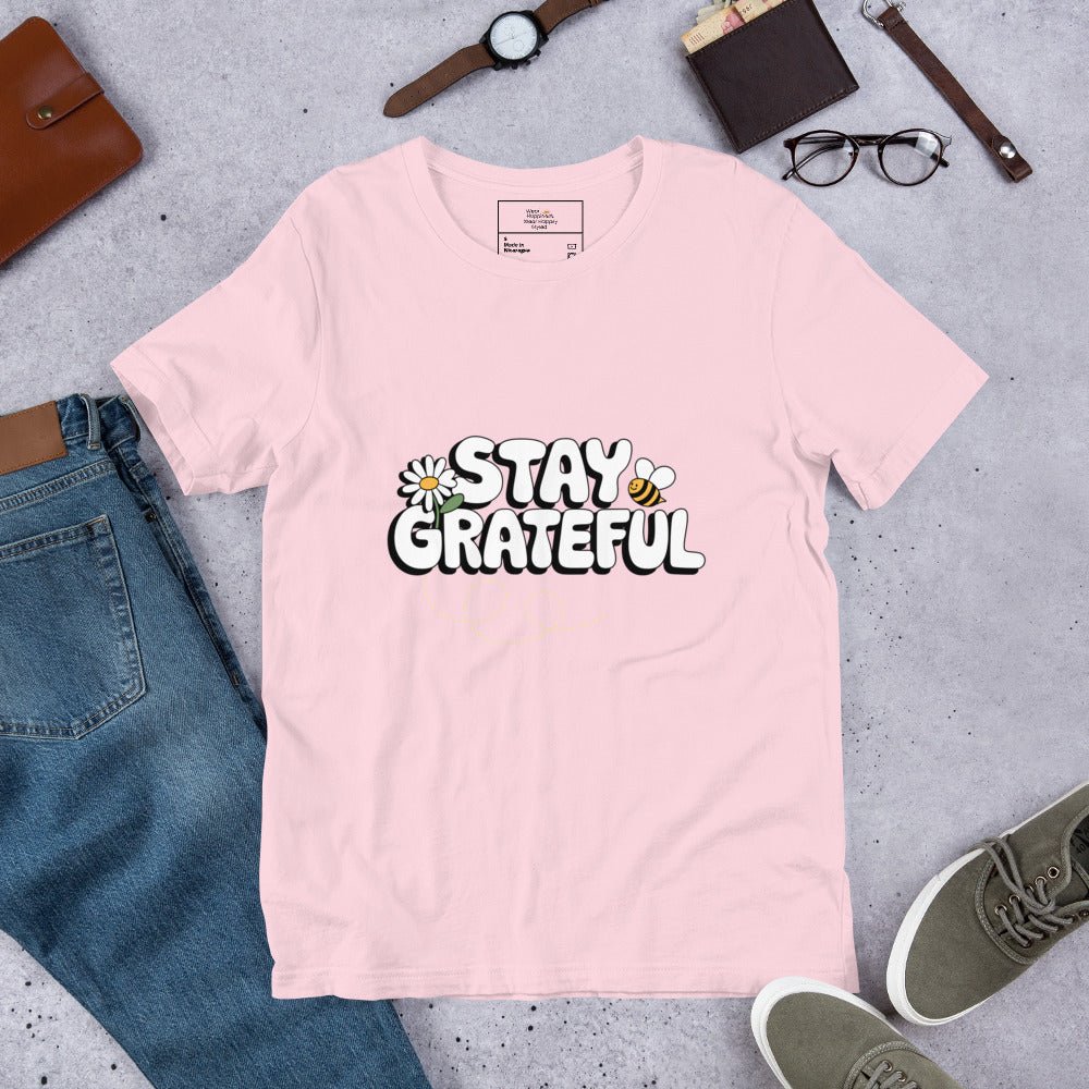 Soft pink t-shirt with “Stay Grateful” text design, featuring daisies and a bee trail, styled flat with jeans, sneakers, glasses, a watch, and leather accessories on a gray background.