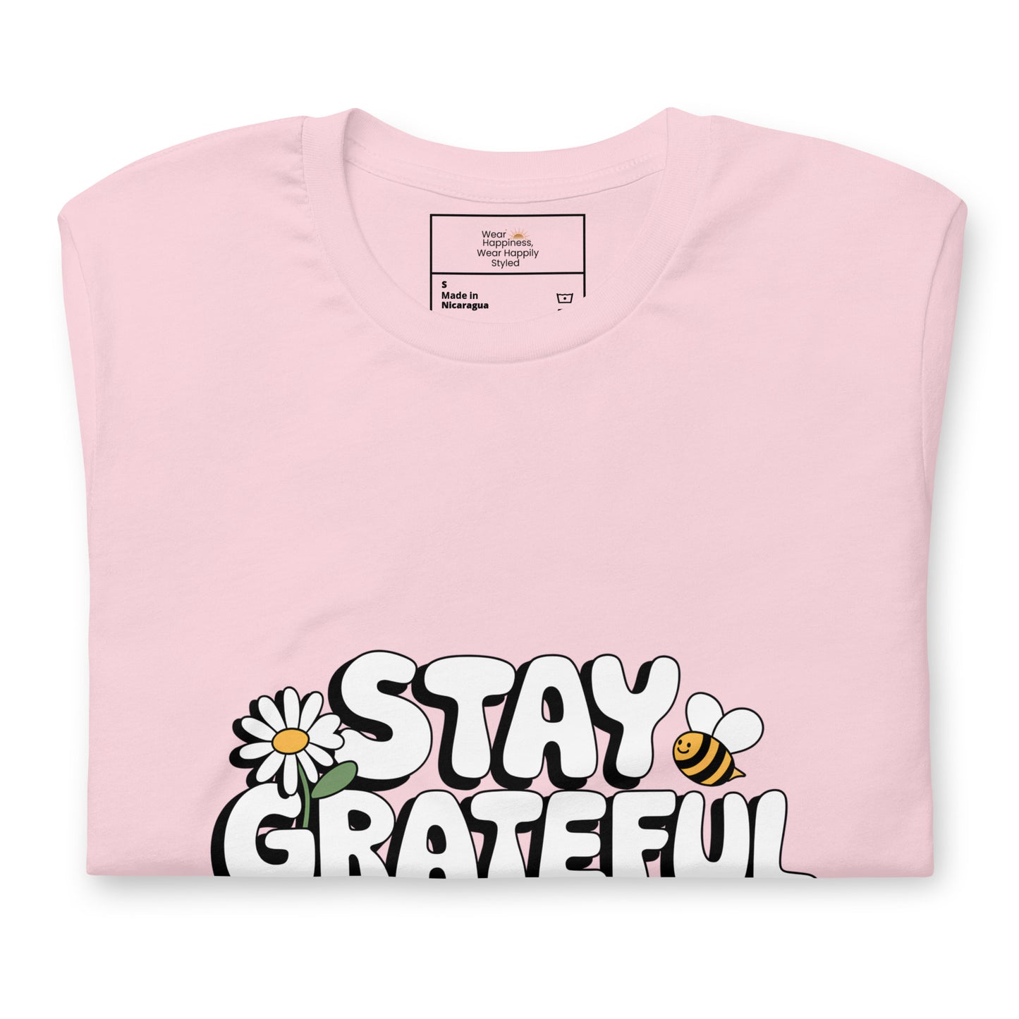 Folded soft pink t-shirt with a bold white “Stay Grateful” design, featuring a daisy flower and a bee illustration, displayed on a white background.