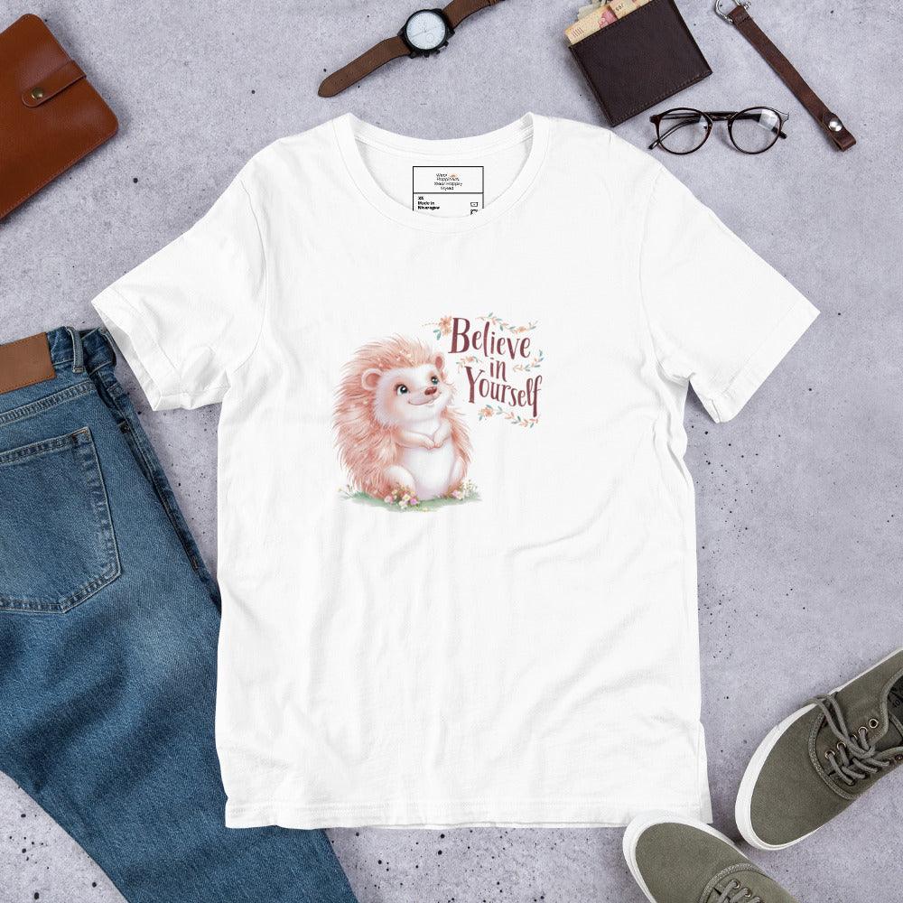 Flat lay of a white t-shirt with a pastel hedgehog and “Believe in Yourself” quote – cozy motivational tee styled with jeans and accessories.