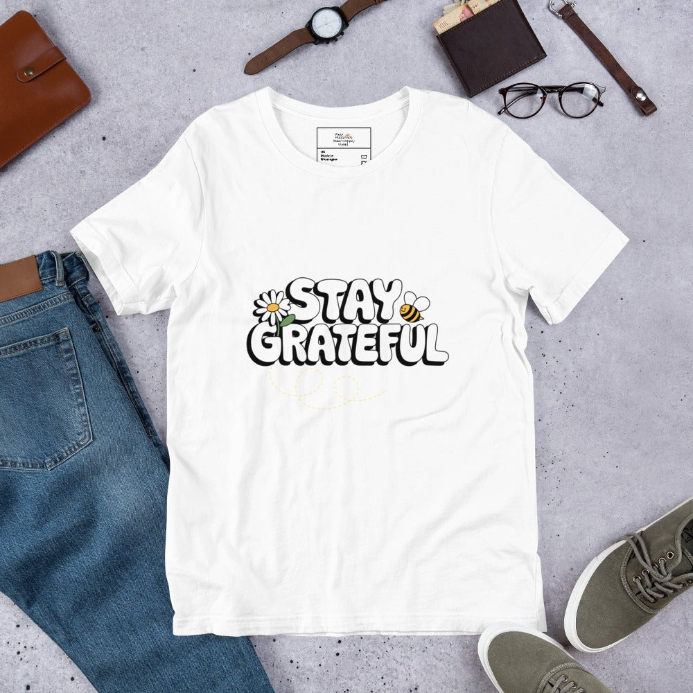 White t-shirt with “Stay Grateful” text design, featuring daisies and a bee trail, styled flat with jeans, sneakers, glasses, a watch, and leather accessories on a gray background.