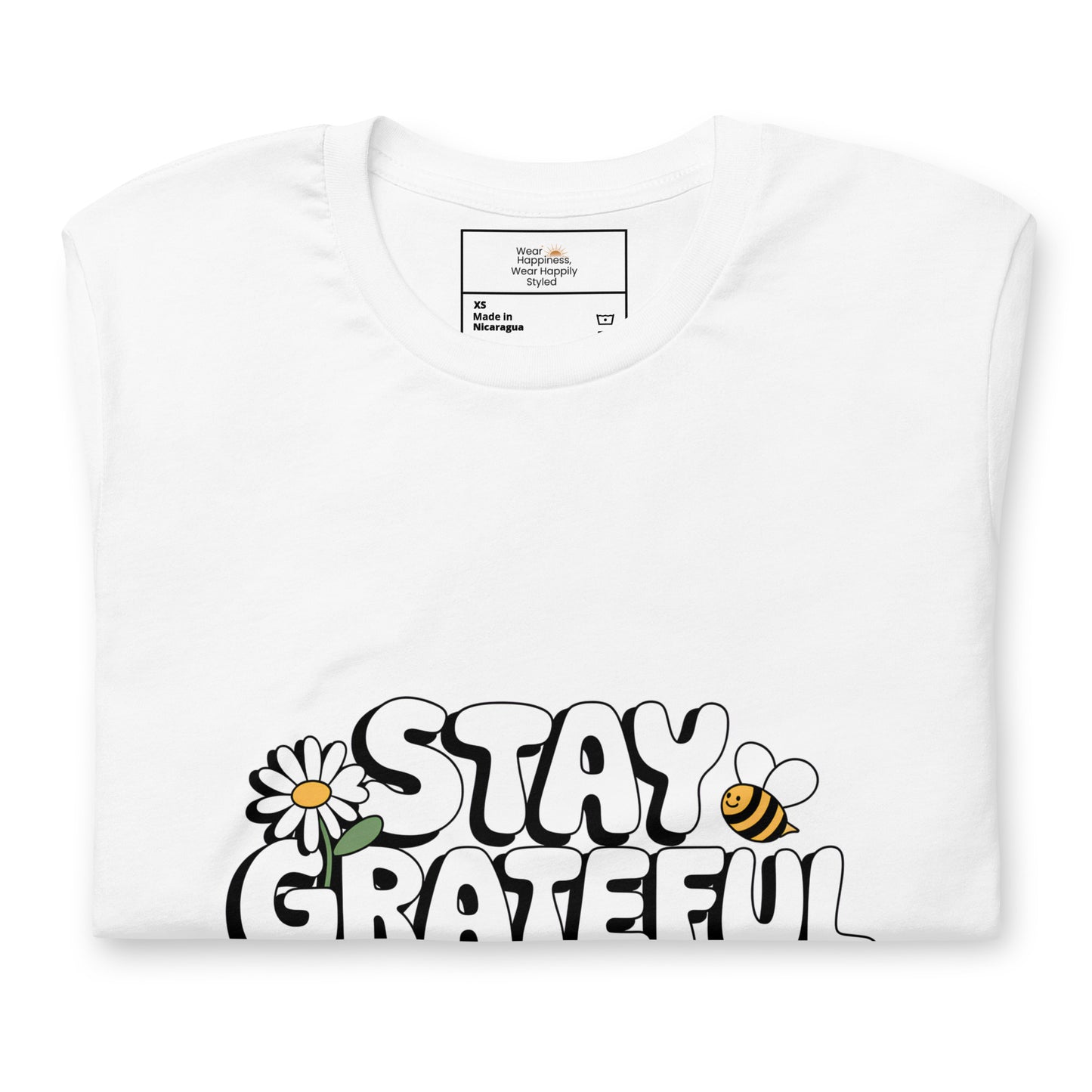 Folded white t-shirt with a bold white “Stay Grateful” design, featuring a daisy flower and a bee illustration, displayed on a white background.