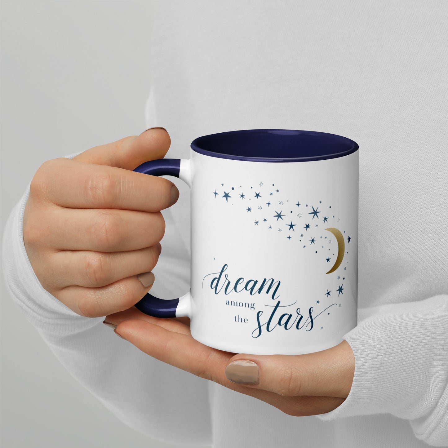 White ceramic mug with dark blue interior and gold moon with stars design, reading “dream among the stars,” held by a person in a white sweater.