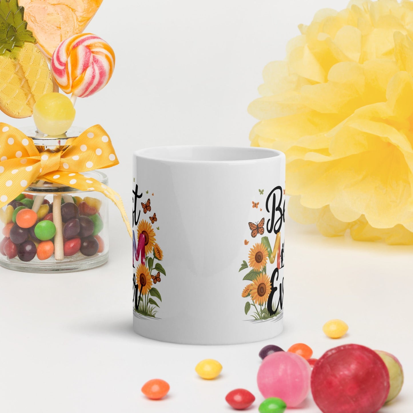 White ceramic mug with floral “Best Mom Ever” design, surrounded by candies and bright yellow decor on a white background.