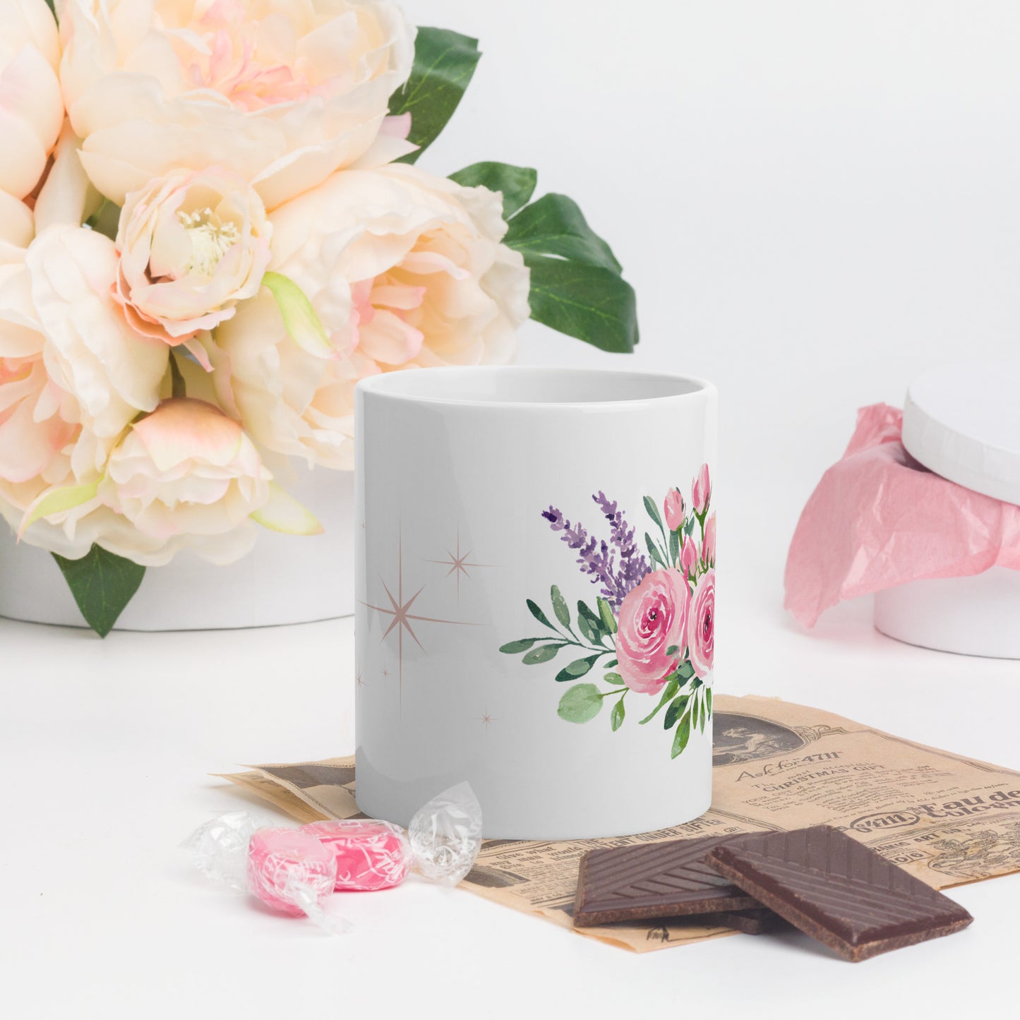White ceramic mug with pink and lavender floral design, styled next to candy, chocolate, and soft pink peonies.
