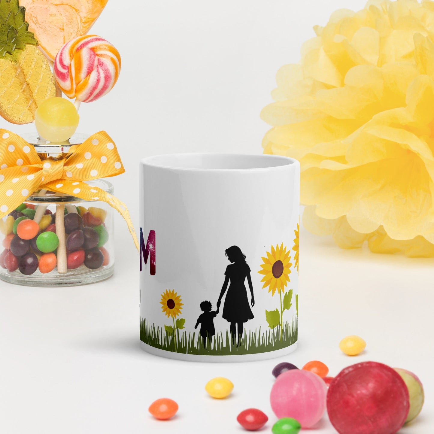 White mug with a silhouette of a mom and child surrounded by sunflowers, placed on a white surface with colorful candies and a yellow tissue flower in the background.