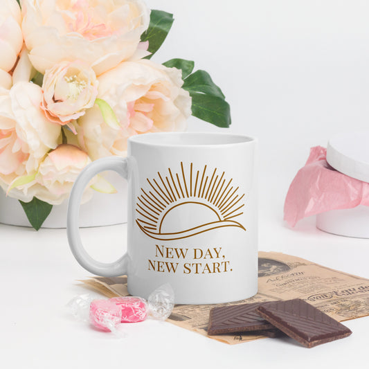 White ceramic mug with gold sun and “New Day, New Start” text, styled with flowers, candy, and chocolate on a white surface.
