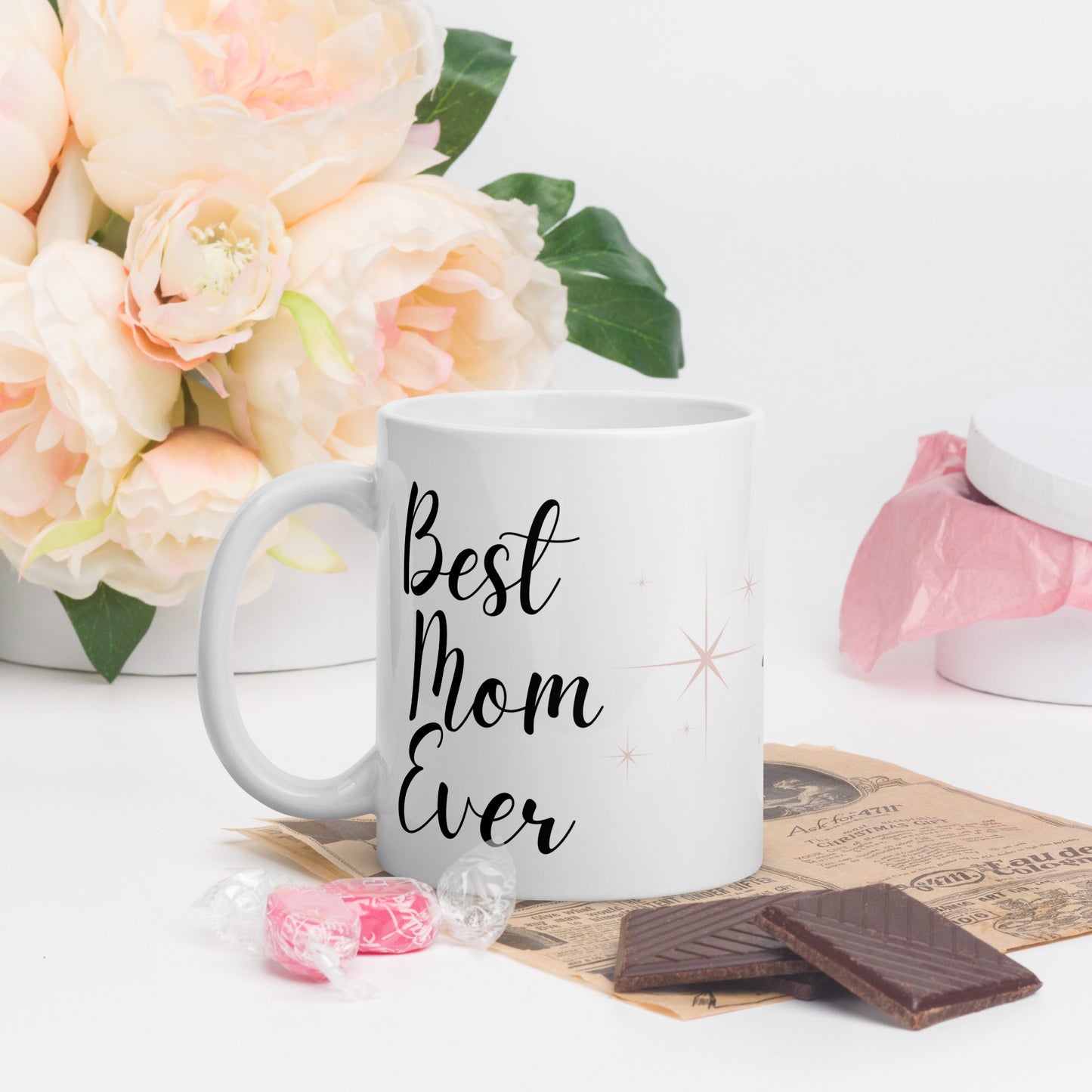 White mug with "Best Mom Ever" text, styled with candy, chocolate, and soft pink flowers in the background.