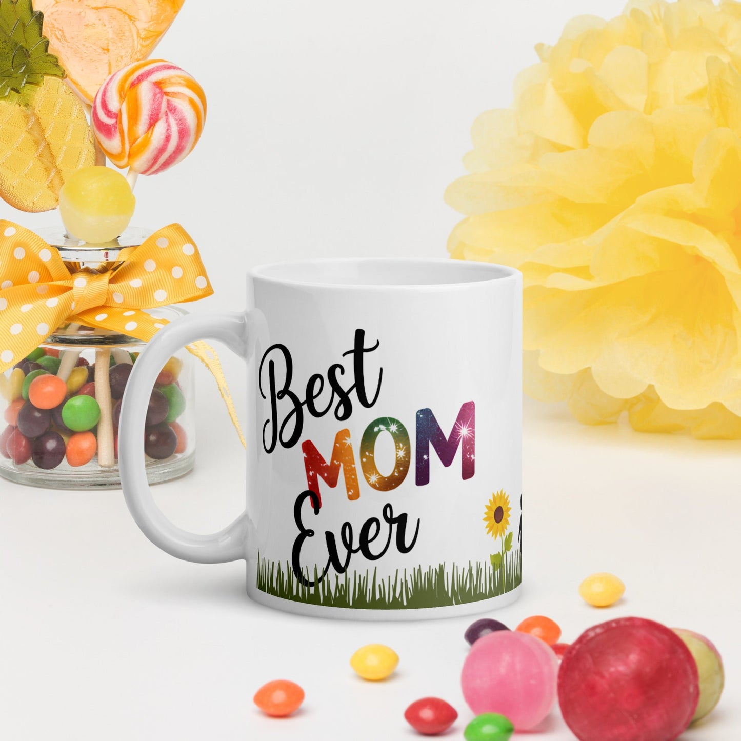 White mug with “Best Mom Ever” rainbow text, handle on the left, surrounded by colorful candy and yellow decor.