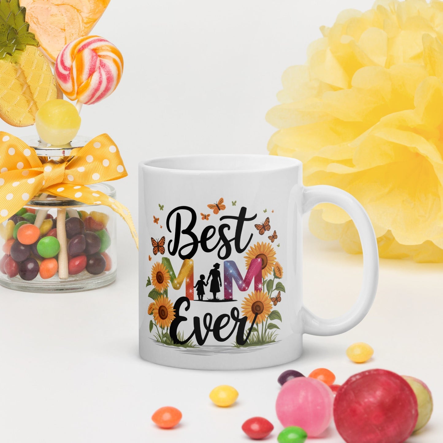 White ceramic mug with floral “Best Mom Ever” design, surrounded by candies and bright yellow decor on a white background.