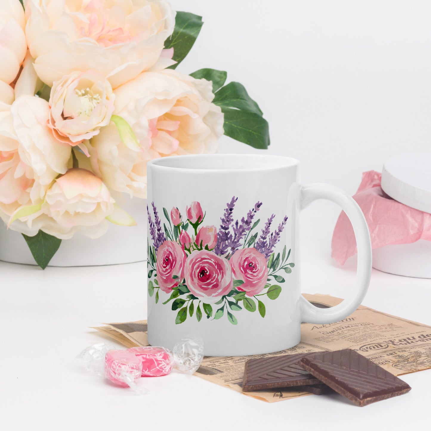 White ceramic mug with pink and lavender floral design, styled next to candy, chocolate, and soft pink peonies.