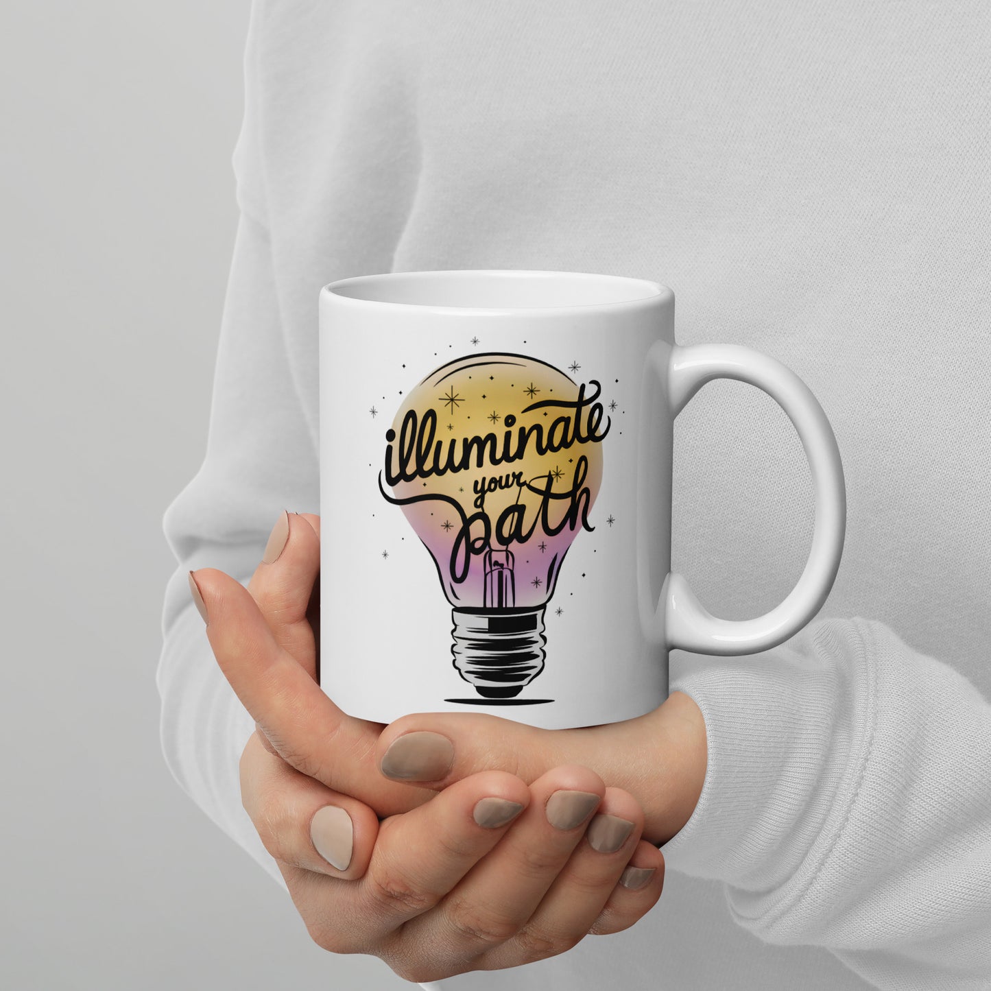 Close-up of woman holding white ceramic mug with “Illuminate Your Path” text and colorful lightbulb design