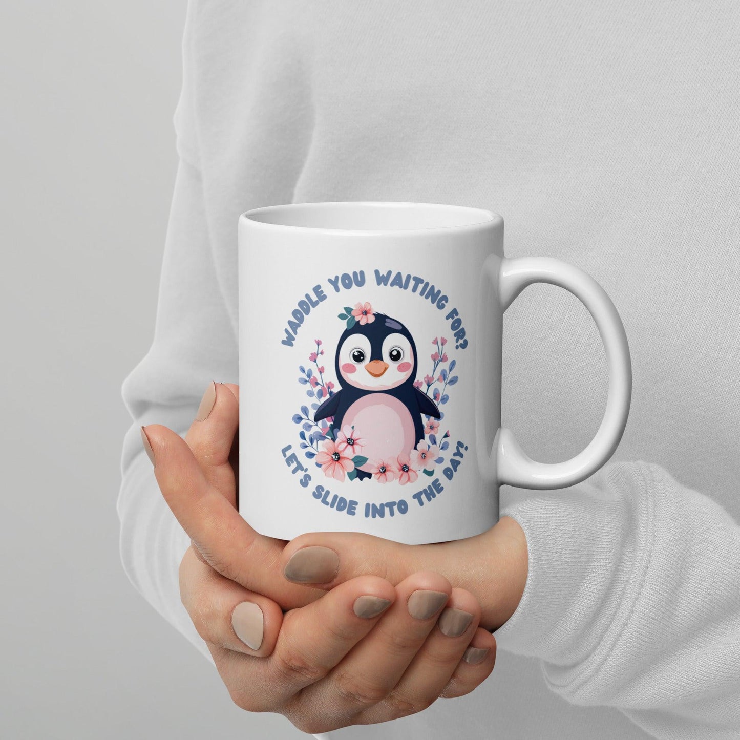 A person wearing a white sweater holding a white mug with a cute penguin illustration and the text “Waddle You Waiting For? Let’s Slide Into the Day!” surrounded by pink and blue flowers.