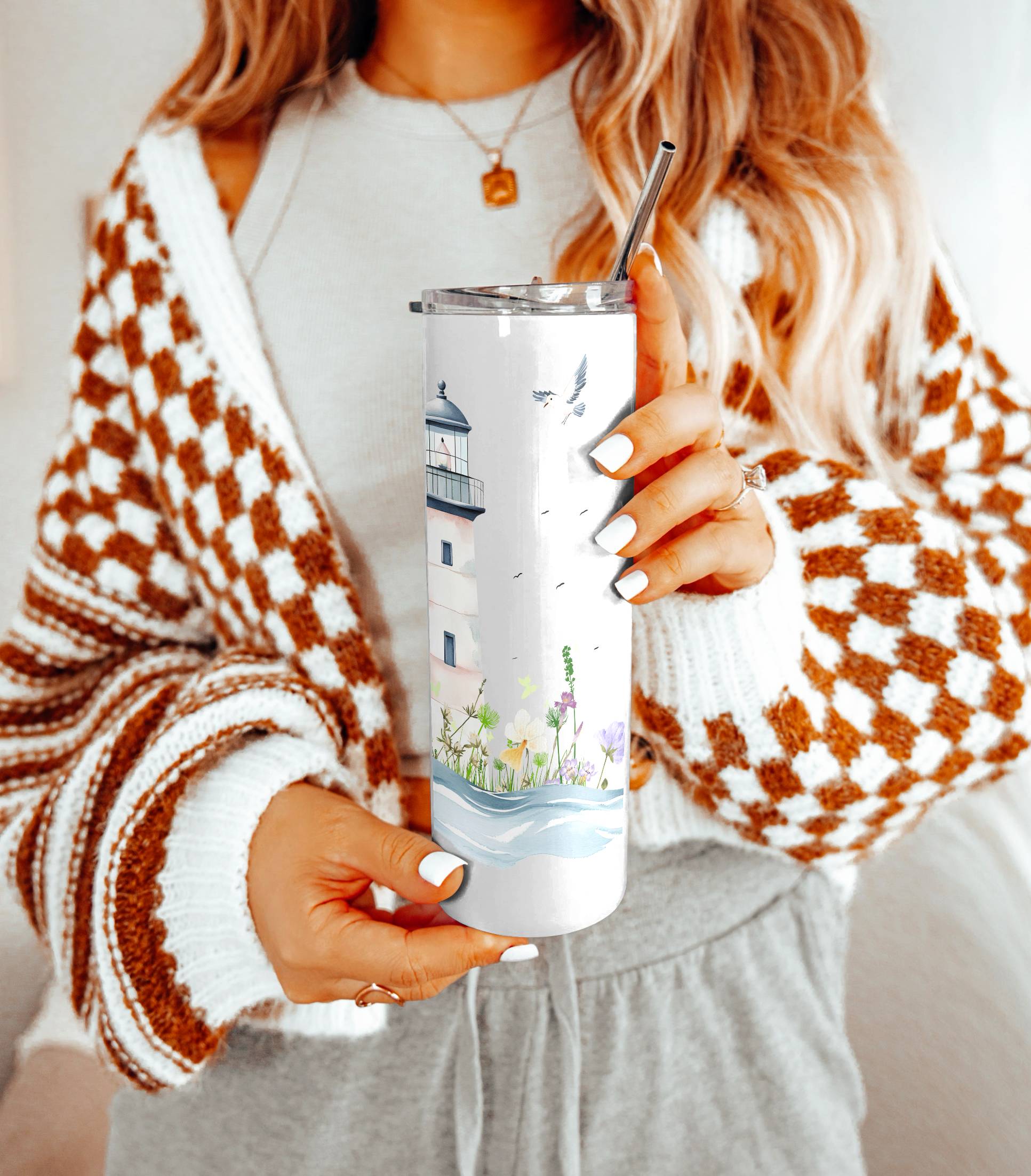 Woman holding a stainless steel tumbler with a pastel lighthouse and floral coastal design, wearing a checkered cardigan and light gray outfit.