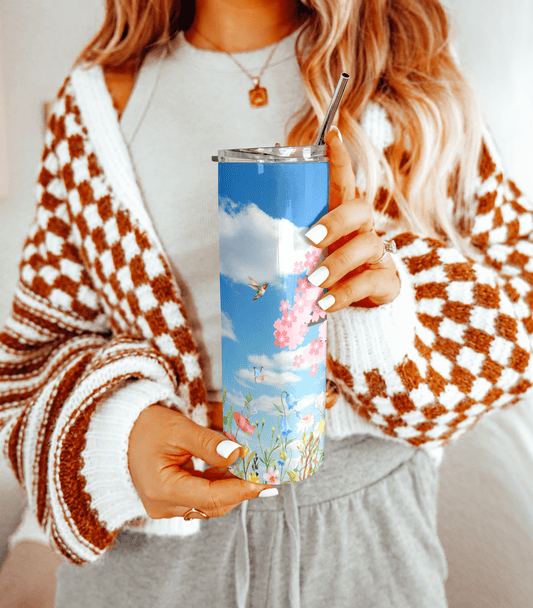 Woman holding a tall stainless steel tumbler with a blue sky and cherry blossom floral design, wearing a checkered brown and white cardigan.