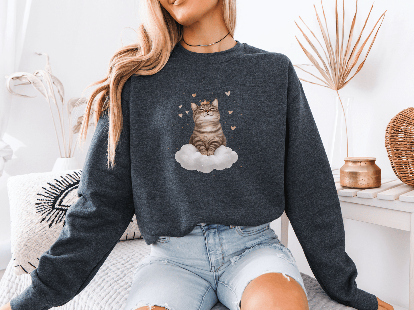 Woman wearing a dark heather sweatshirt featuring a majestic tabby cat sitting on a fluffy cloud, surrounded by soft pink hearts 