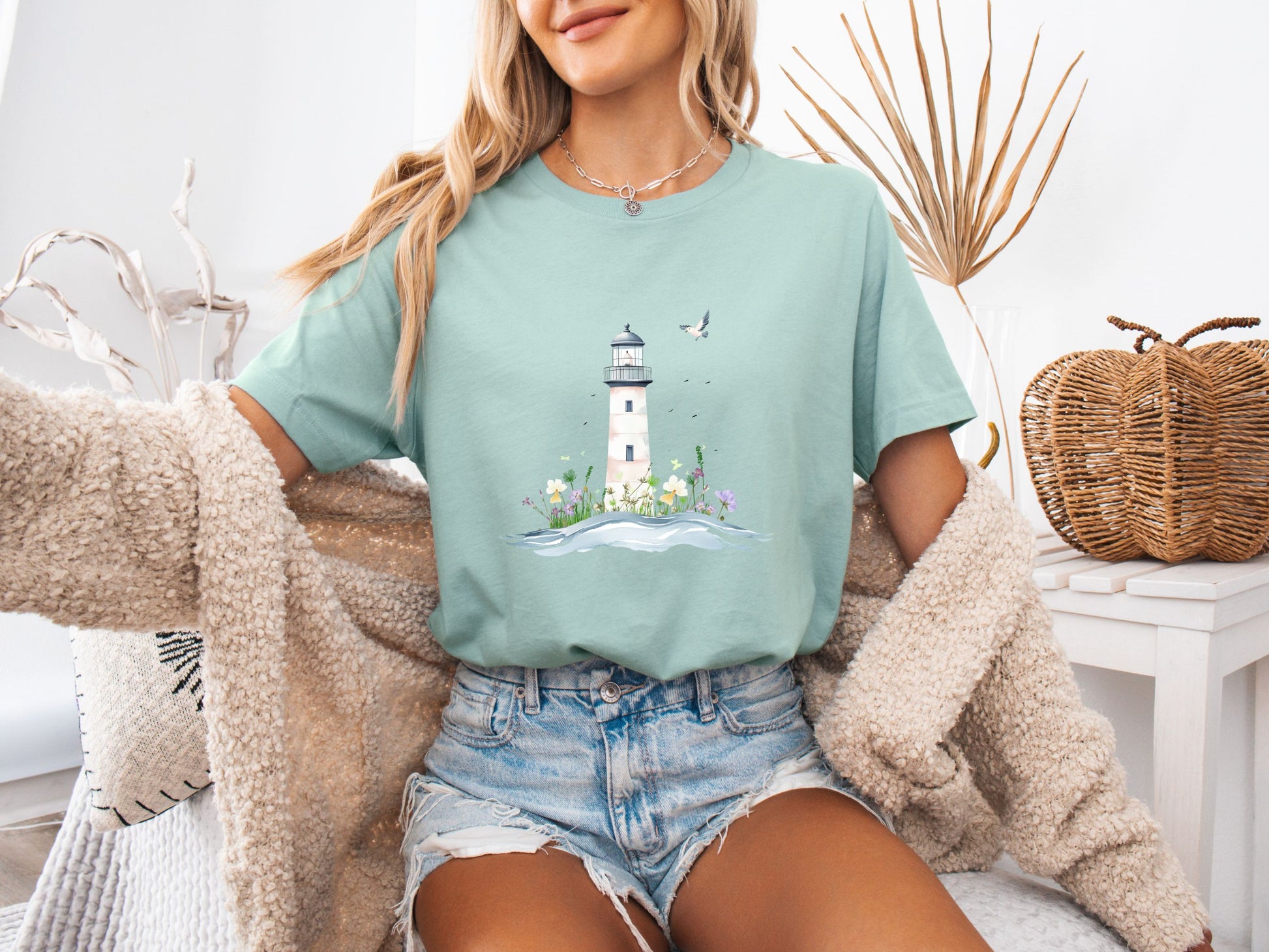 Woman sitting on a couch wearing a dusty blue t-shirt with a whimsical lighthouse and floral design, paired with denim shorts and a cozy knit cardigan, in a calm and stylish home setting.