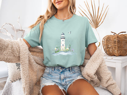 Woman sitting on a couch wearing a dusty blue t-shirt with a whimsical lighthouse and floral design, paired with denim shorts and a cozy knit cardigan, in a calm and stylish home setting.