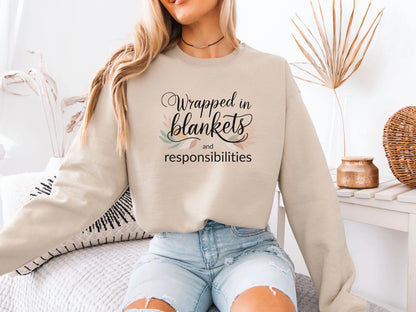 Person wearing a beige sweatshirt with text "Wrapped in blankets and responsibilities", sitting on a couch holding a mug.