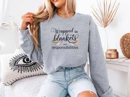 Person wearing a grey sweatshirt with text "Wrapped in blankets and responsibilities", sitting on a couch holding a mug.