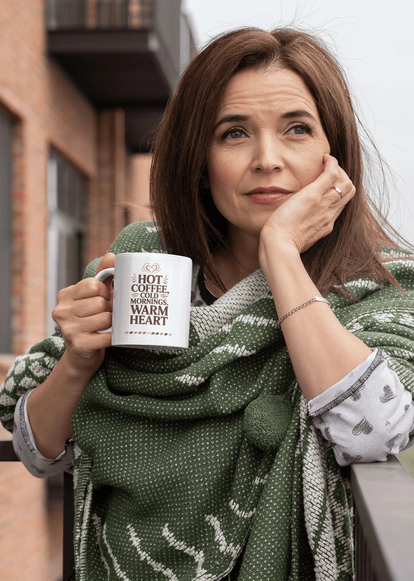 Woman holding a mug with text "Hot coffee cold mornings warm heart" sitting outdoors.