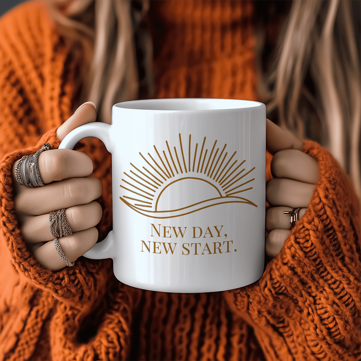 White mug with gold sun illustration and “New Day, New Start” quote, held by woman in cozy orange sweater.