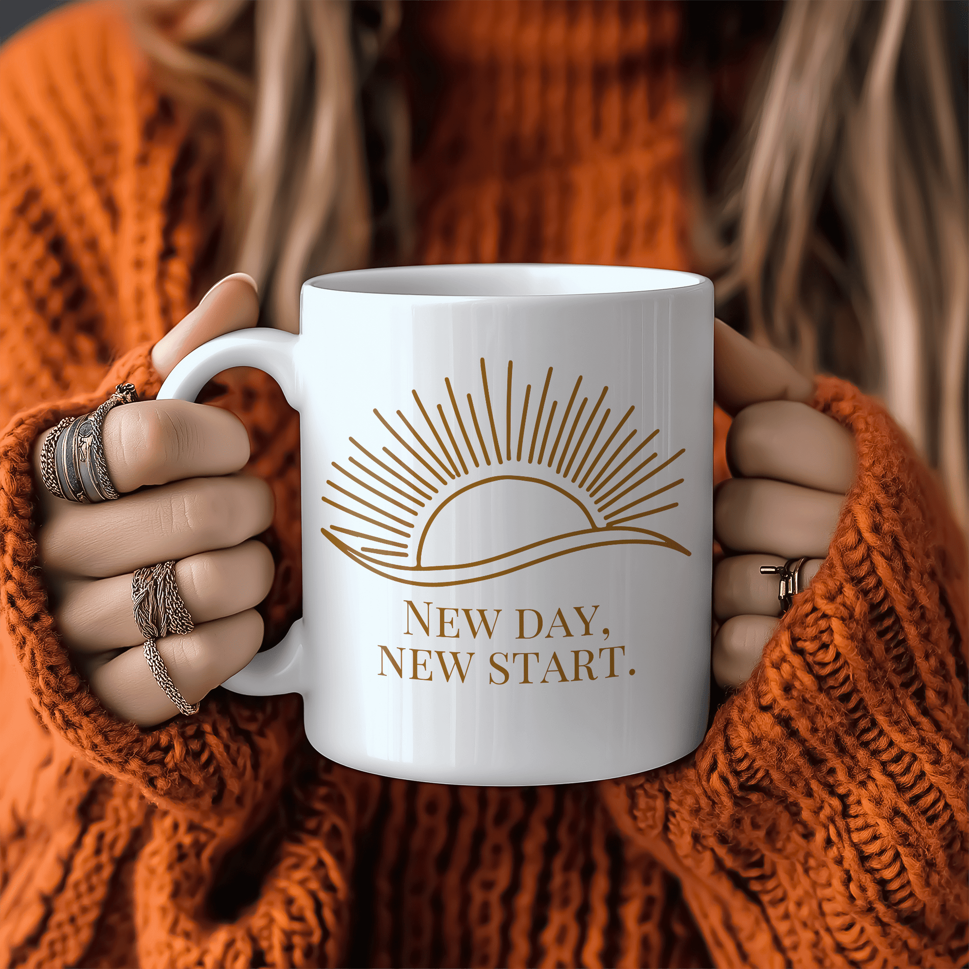 White mug with gold sun illustration and “New Day, New Start” quote, held by woman in cozy orange sweater.