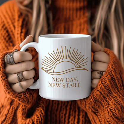 White mug with gold sun illustration and “New Day, New Start” quote, held by woman in cozy orange sweater.