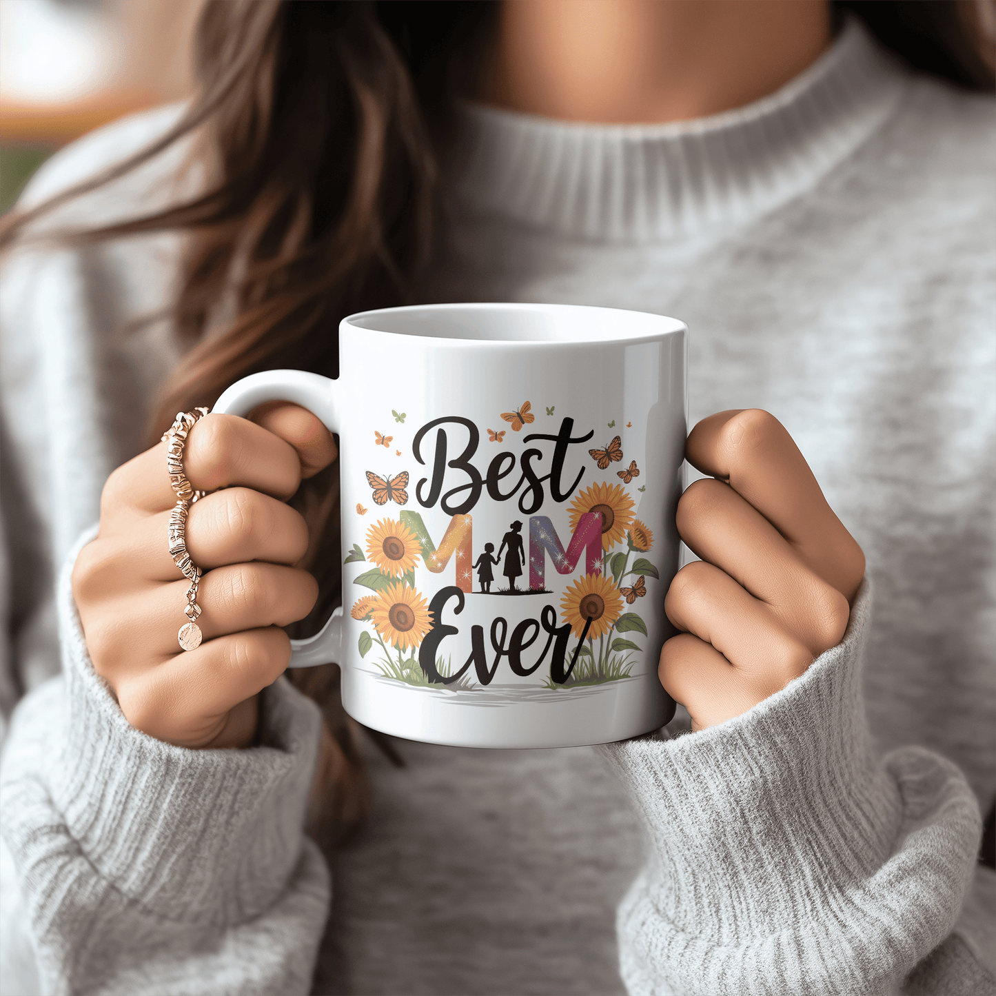 Woman holding a white mug with “Best Mom Ever” text, decorated with sunflowers and butterflies, wearing a cozy gray sweater.