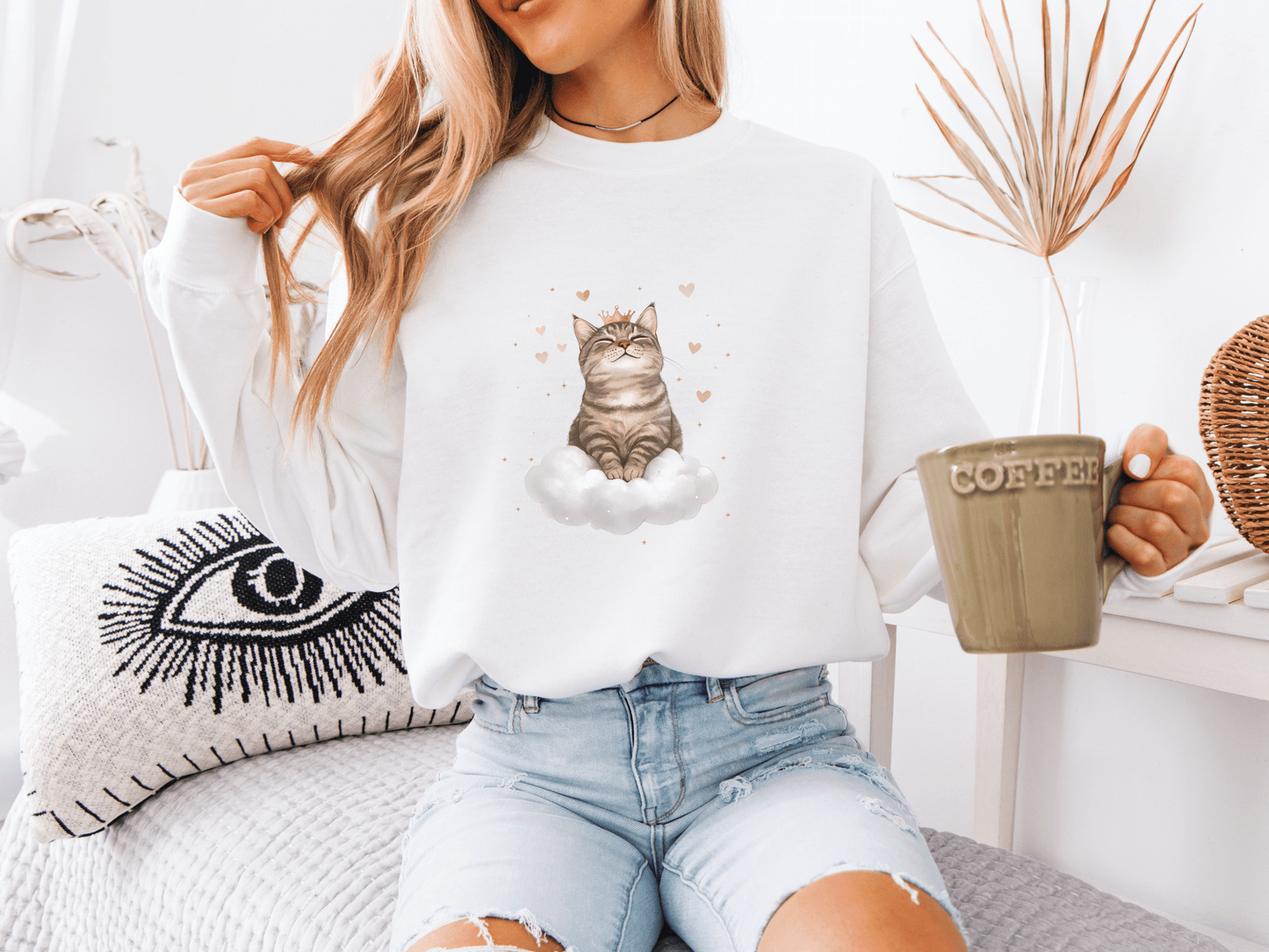 Woman holding a green coffee mug and wearing a white sweatshirt featuring a regal tabby cat sitting on a cloud
