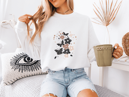 Woman wearing a white sweatshirt featuring elegant black and blush butterflies and flowers, holding a coffee mug – a soft and stylish gift for moms who love whimsical bloom designs.
