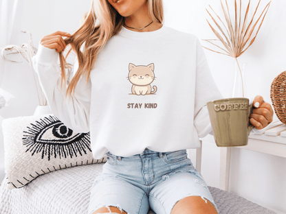 Woman sitting on a bed, wearing a white sweatshirt with a cute cat illustration and the words “Stay Kind,” holding a large green coffee mug in a bright, cozy room.