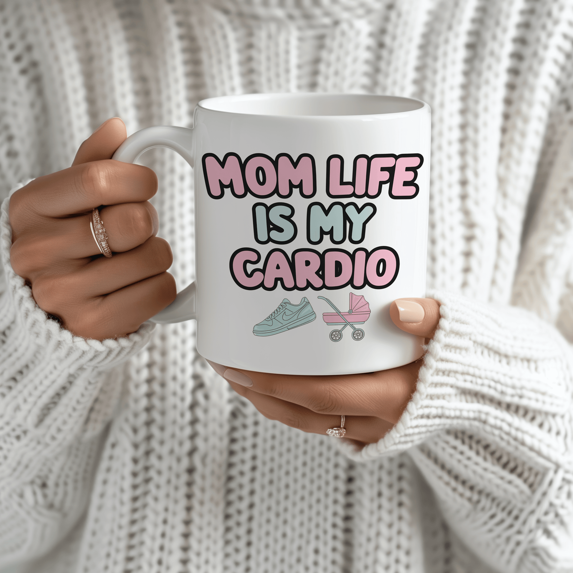White mug with “Mom Life Is My Cardio” in bold pink and green letters, held by a woman in a cozy white sweater.