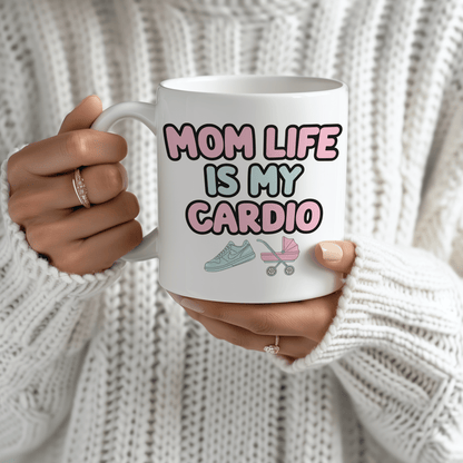 White mug with “Mom Life Is My Cardio” in bold pink and green letters, held by a woman in a cozy white sweater.
