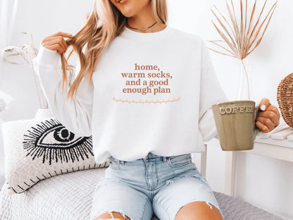 Person wearing a white sweatshirt with text "home, warm socks, and a good enough plan", holding a mug, sitting on a couch.