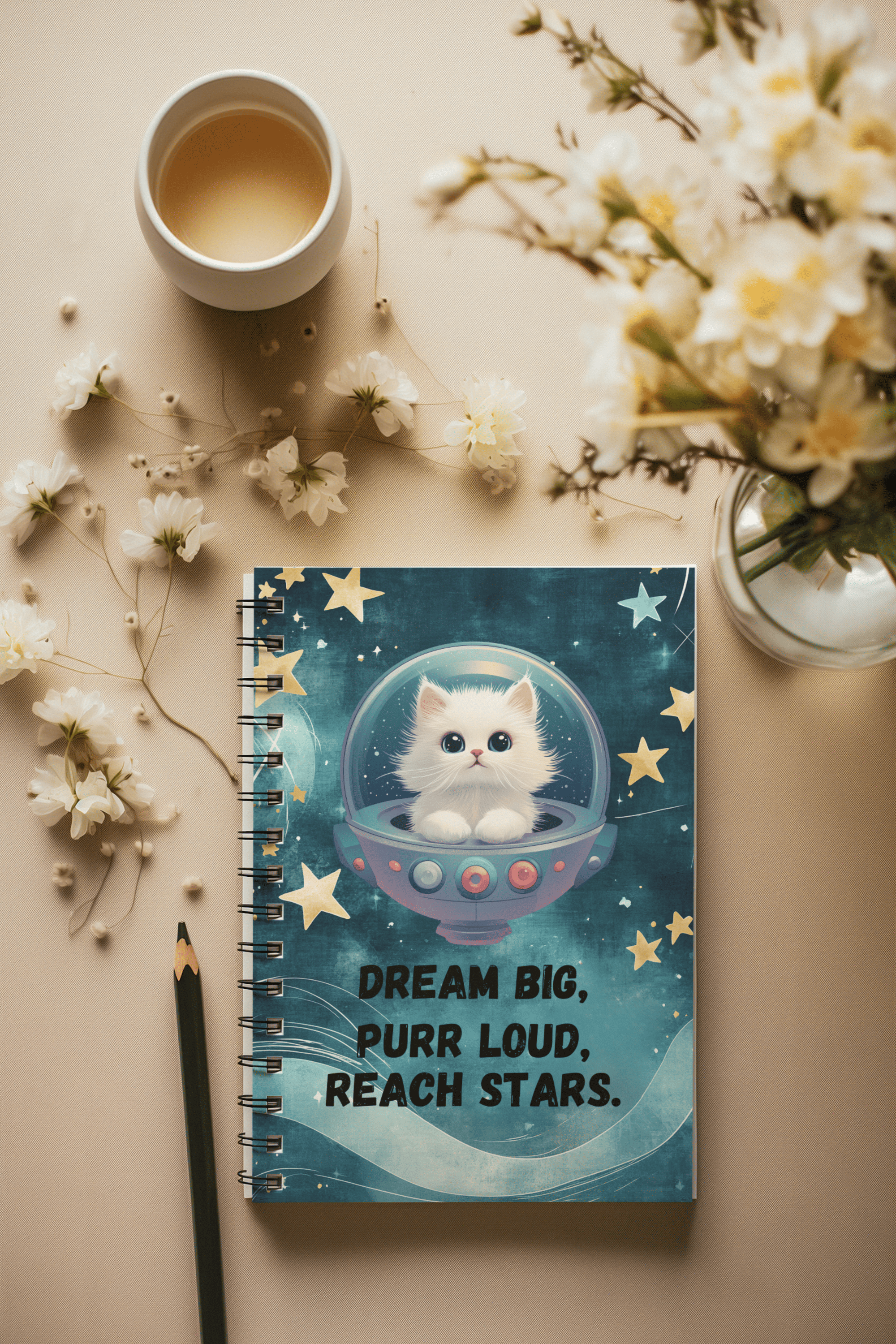 Spiral notebook with a blue galaxy background, featuring a cute white cat in a space helmet and the motivational phrase “Dream Big, Purr Loud, Reach Stars.” The notebook is placed on a beige surface with white flowers, a pencil, and a cup of tea.