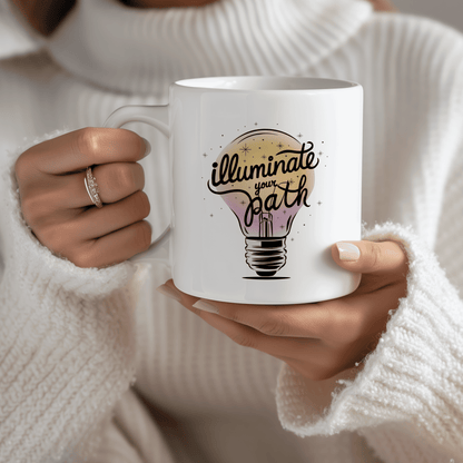 Woman in cozy white sweater holding a white ceramic mug with hand-lettered design that says "Illuminate Your Path" over a glowing lightbulb illustration. Moms