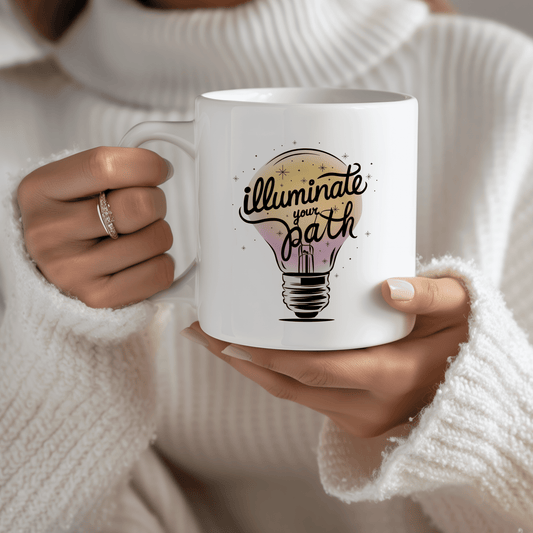Woman in cozy white sweater holding a white ceramic mug with hand-lettered design that says "Illuminate Your Path" over a glowing lightbulb illustration. Moms