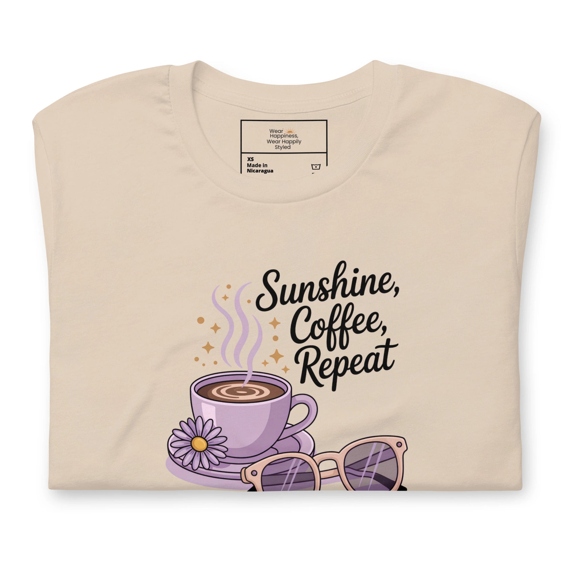 Folded cream t-shirt showing close-up of “Sunshine, Coffee, Repeat” graphic with pastel latte art, sunglasses and sparkles