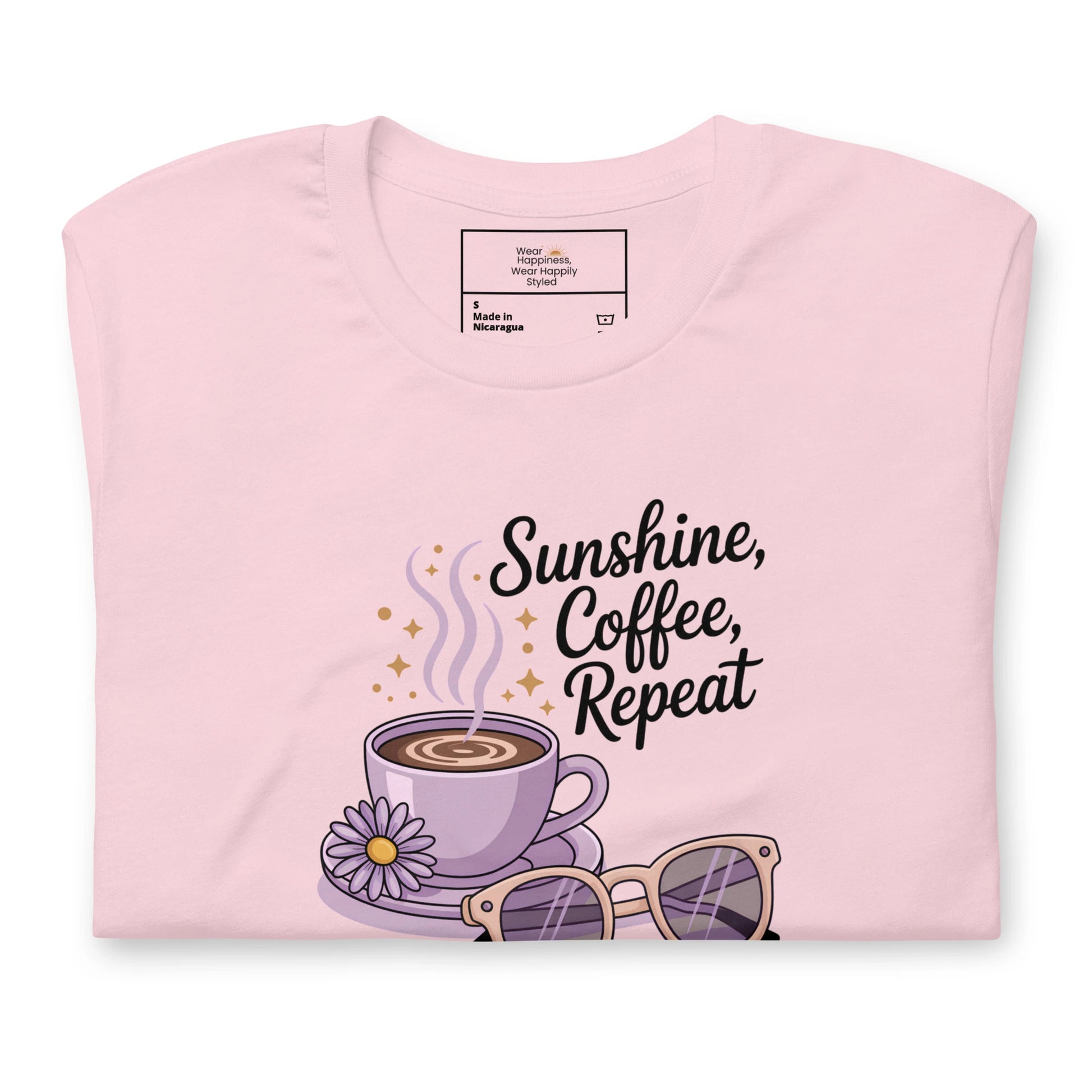 Folded pink t-shirt showing close-up of “Sunshine, Coffee, Repeat” graphic with pastel latte art, sunglasses and sparkles