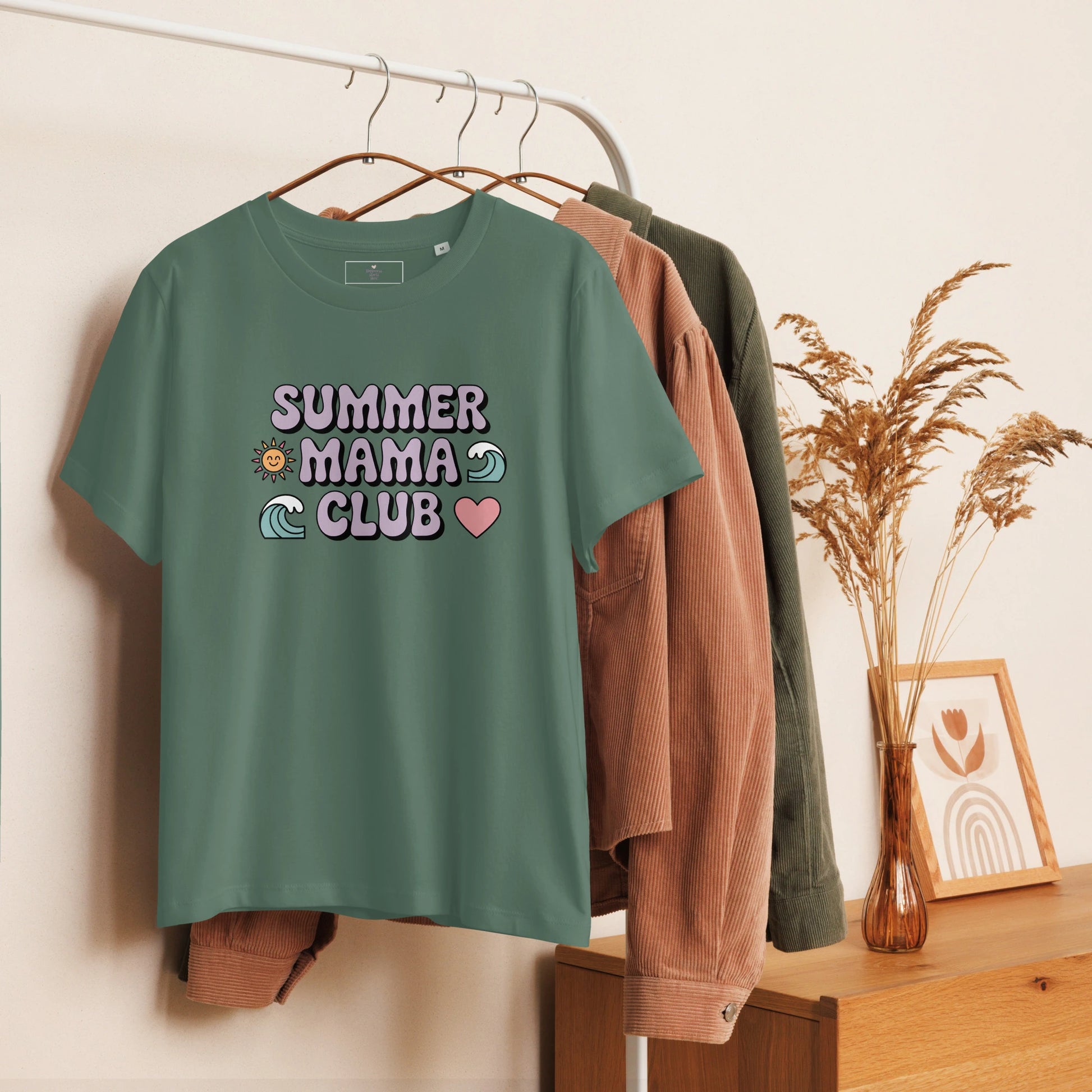 Green t-shirt with pastel "Summer Mama Club" design hanging on a white rack with cozy boho decor in the background.