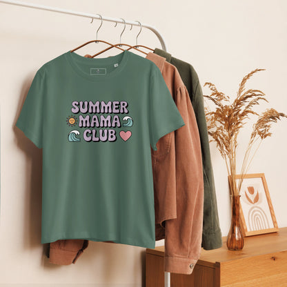 Green t-shirt with pastel "Summer Mama Club" design hanging on a white rack with cozy boho decor in the background.