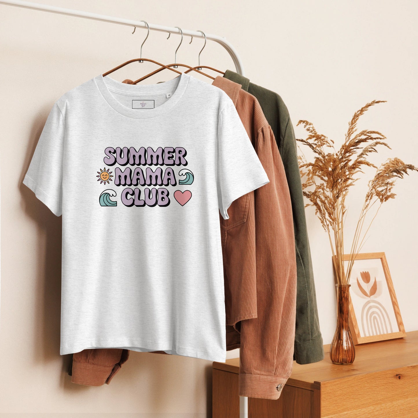 Light grey t-shirt with pastel "Summer Mama Club" design hanging on a white rack with cozy boho decor in the background.