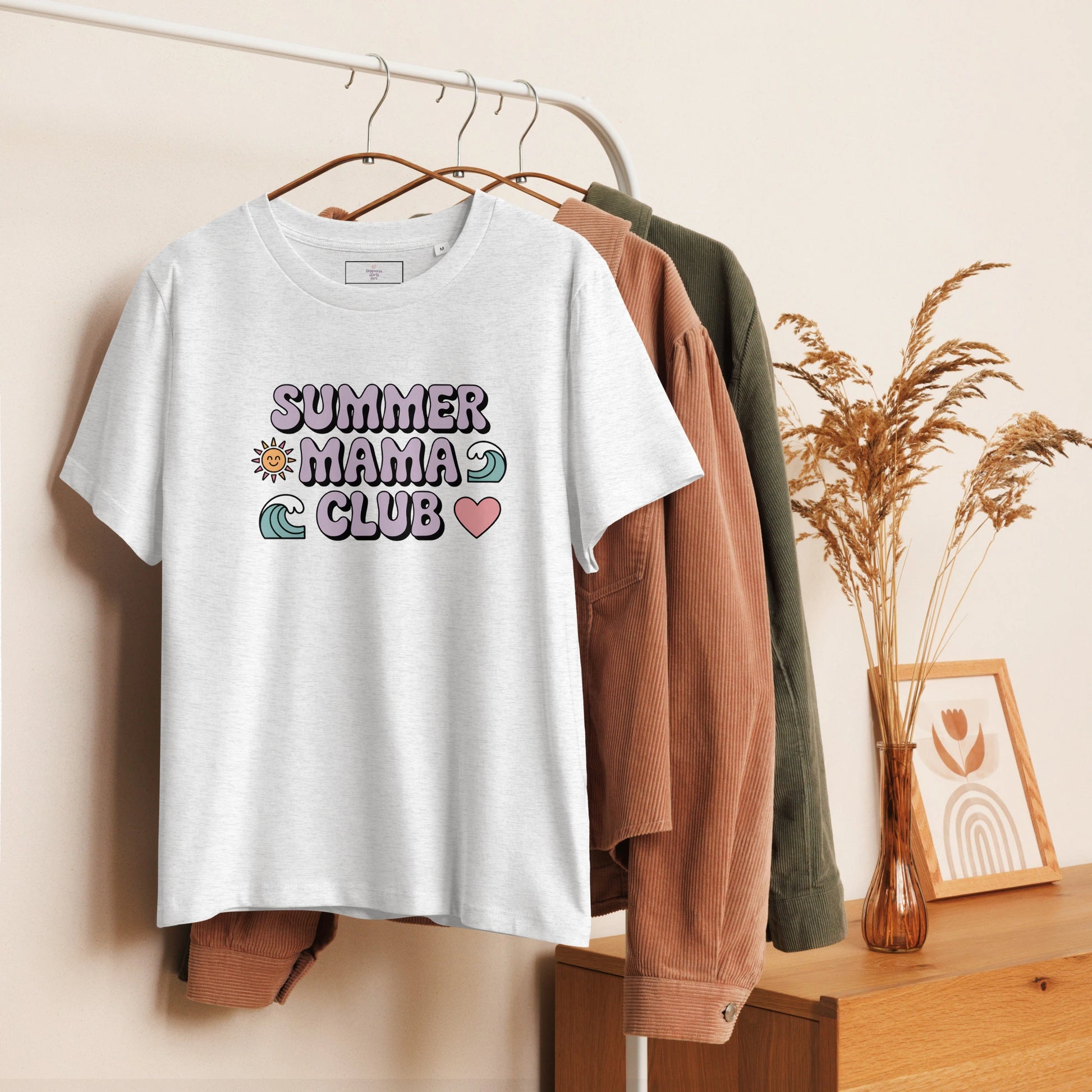 Light grey t-shirt with pastel "Summer Mama Club" design hanging on a white rack with cozy boho decor in the background.