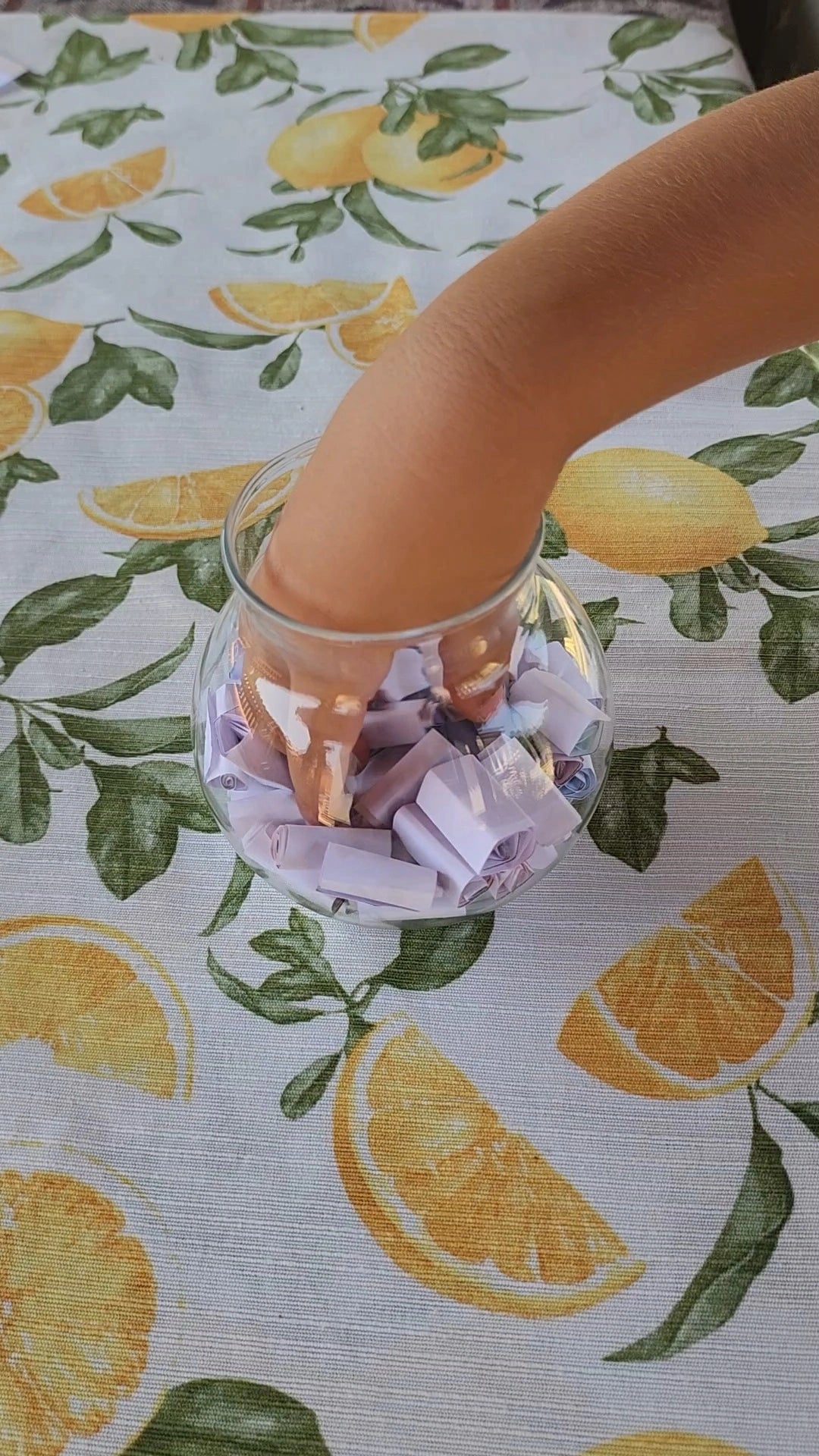 A child’s hand reaching into a glass jar filled with rolled-up summer activity strips on a citrus-themed tablecloth.